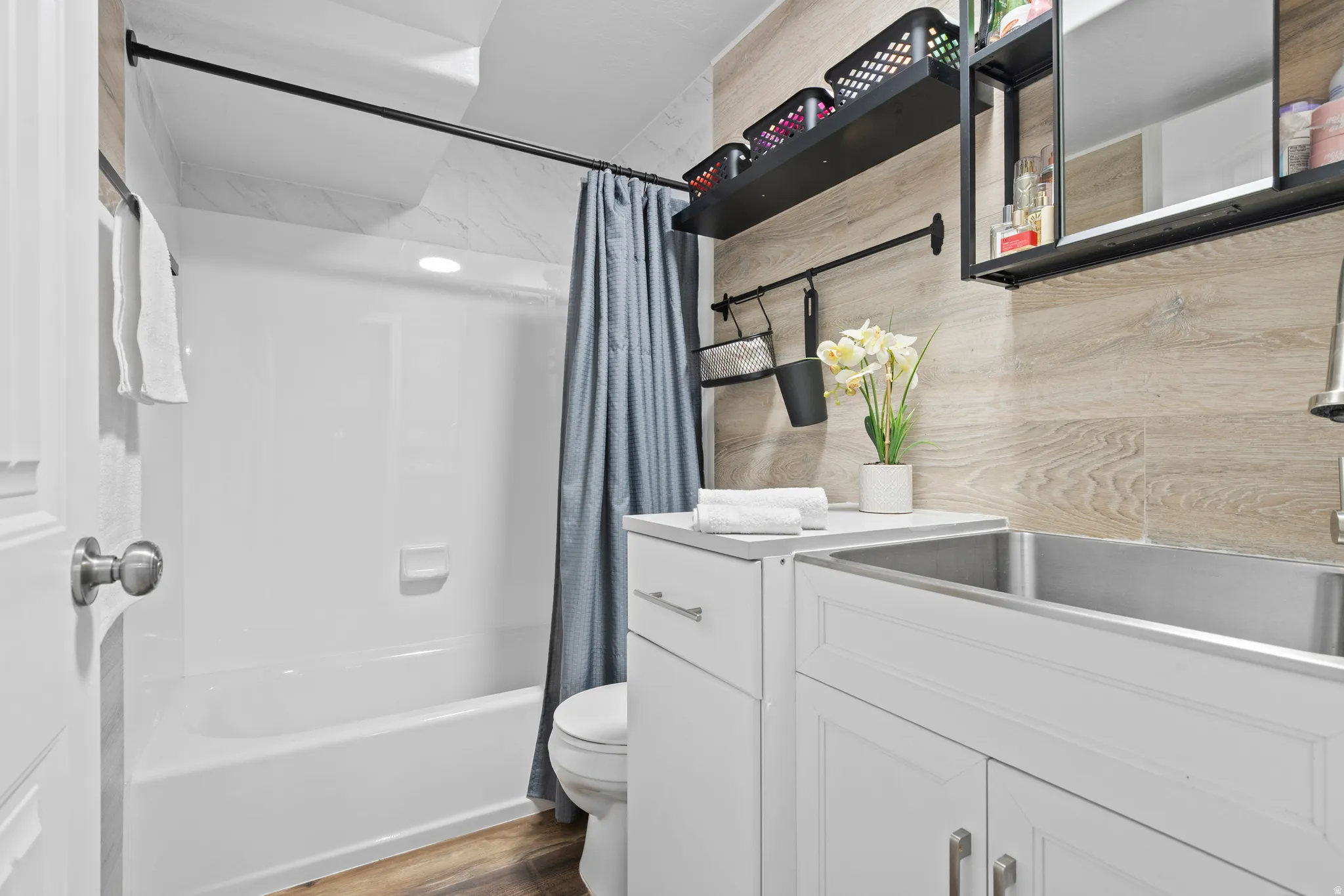 Full bathroom featuring a white tub/shower combination, wood-finish wall paneling, a stainless steel utility sink, white cabinetry, and wood-finish flooring