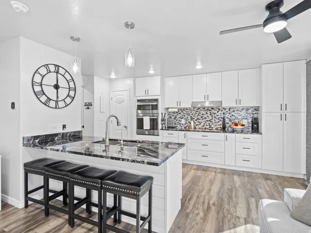 Bright kitchen featuring a central island with a sink and modern faucet, white cabinetry with dark hardware, a mosaic tile backsplash, and built-in double ovens