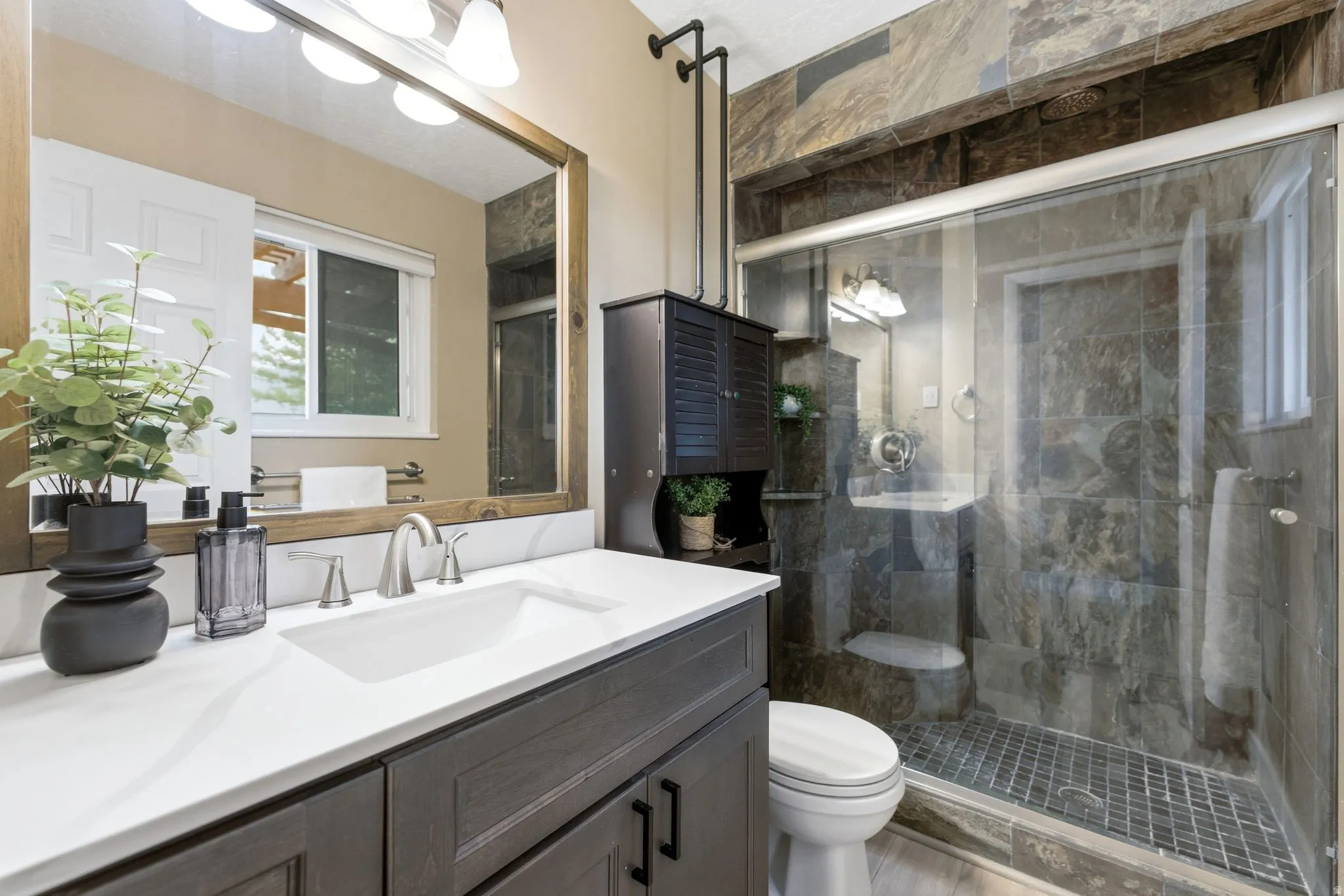 Primary bathroom featuring a dark wood-finish vanity with a white countertop and undermount sink, brushed nickel faucet, large mirror with a wood frame, and a glass-enclosed shower with slate-style tile walls and a mosaic tile floor