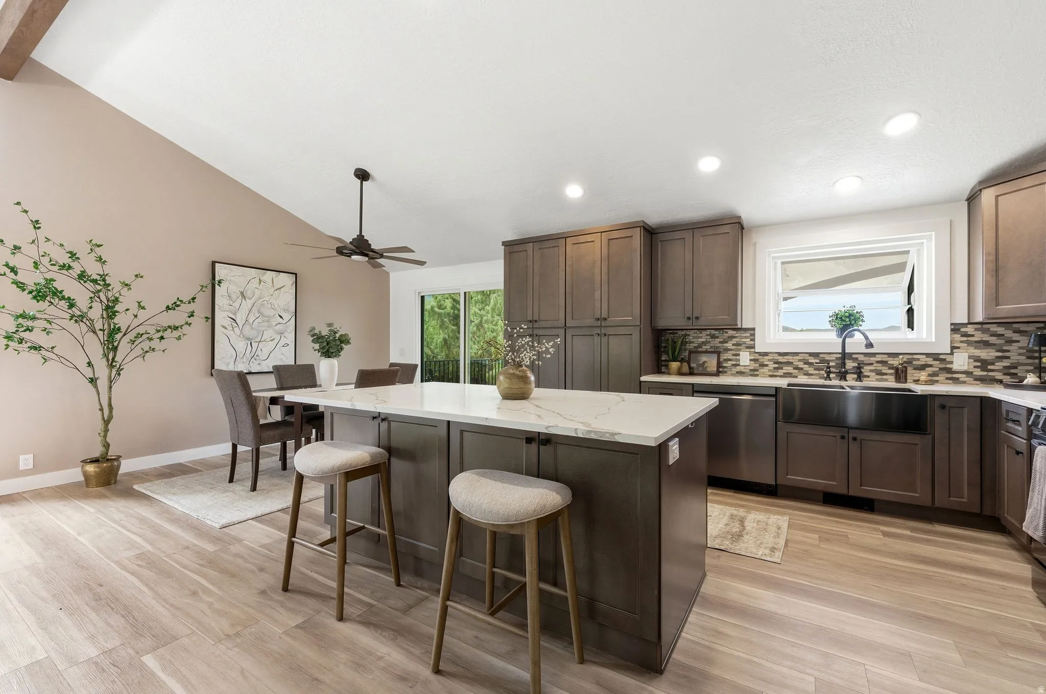 Open-concept kitchen and dining area featuring a vaulted ceiling with exposed beam, wood-finish flooring, a kitchen island with seating, dark wood cabinetry, and stainless-steel appliances
