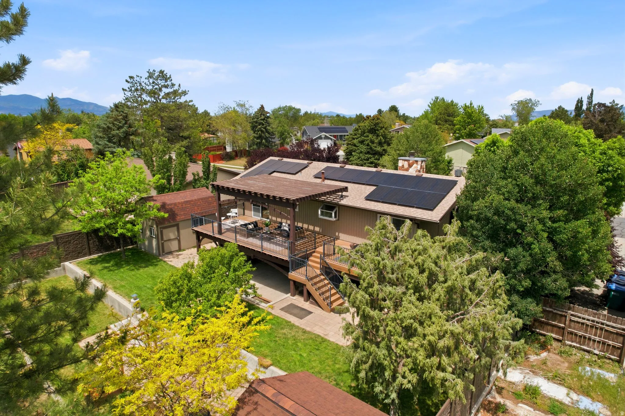 Expansive backyard featuring a multi-level deck with a pergola, a manicured lawn, a detached storage shed, solar panels on the roof, and mature perimeter trees