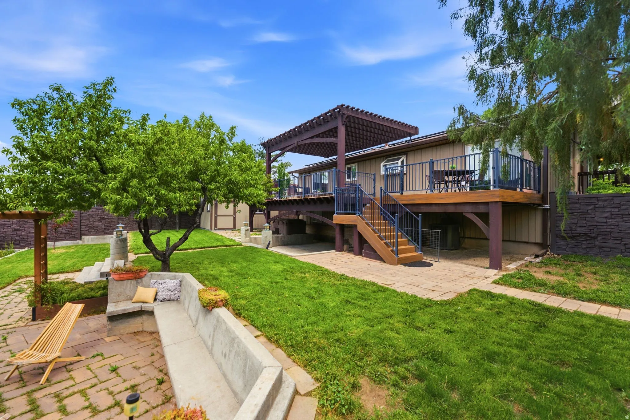 Tiered backyard featuring a spacious deck with a pergola, lower patio area, built-in concrete seating, and lush green lawn