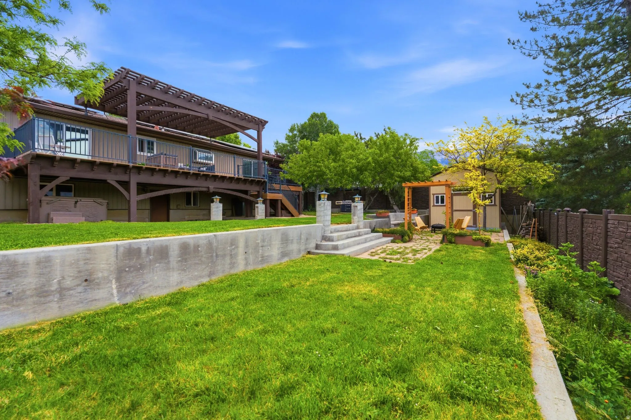 Expansive backyard featuring a large elevated deck with a pergola, a manicured lawn, concrete retaining wall, and a separate structure with a small pergola
