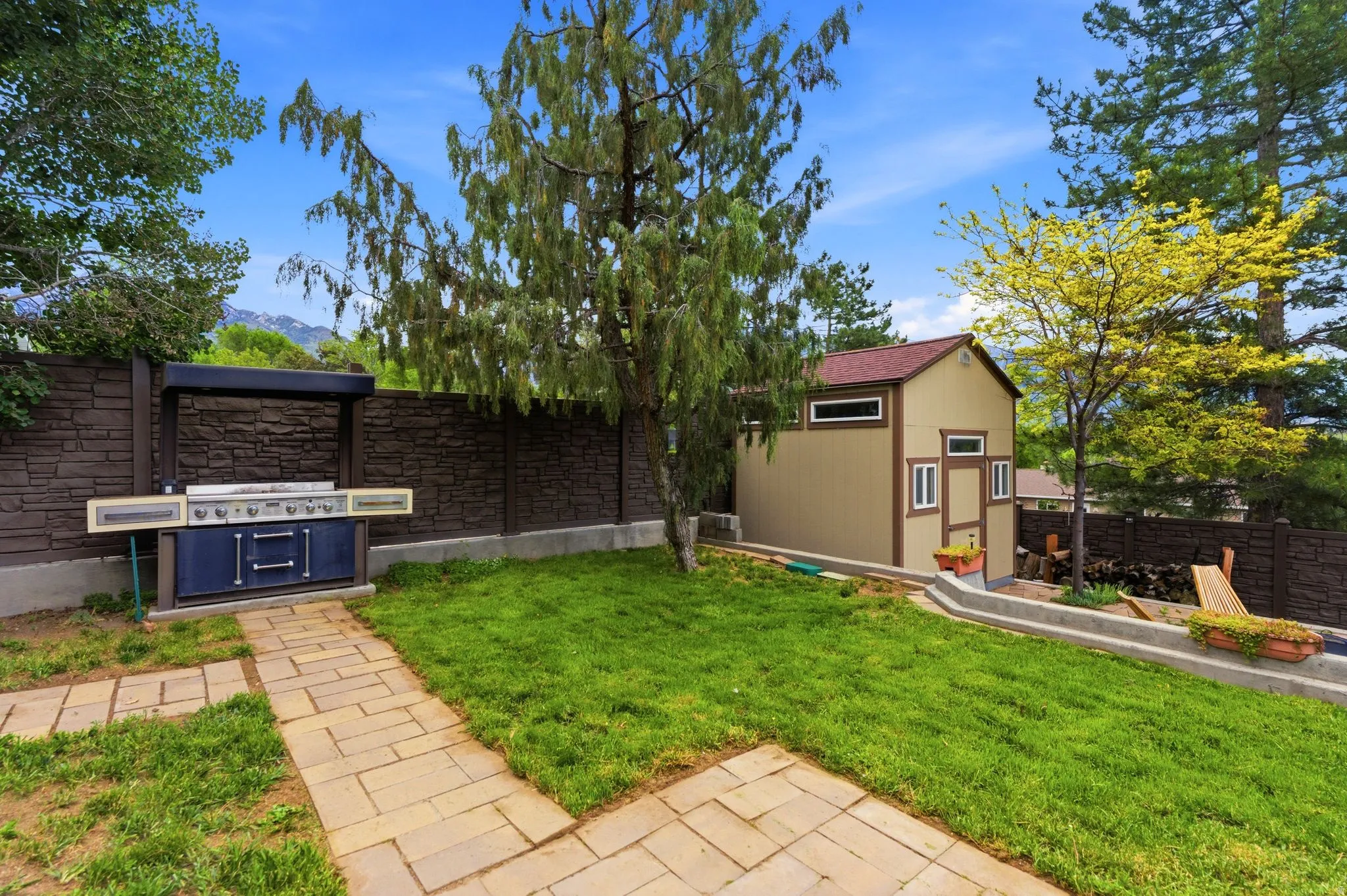 Expansive grassy yard featuring a stone paver pathway, built-in outdoor grill station, mature trees, a detached storage shed with a gabled roof, and a stone retaining wall