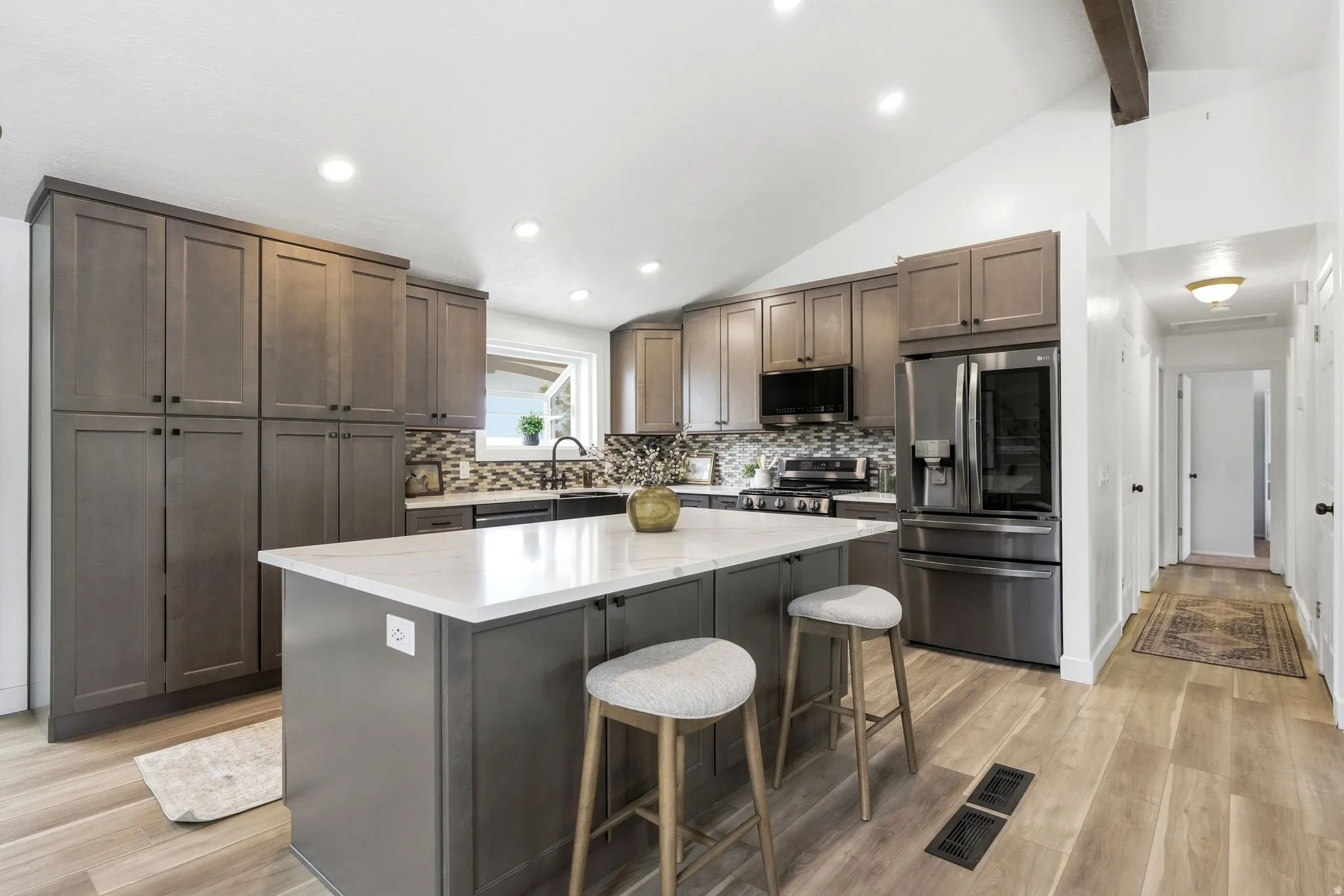 Open-concept kitchen featuring an oversized island with a white solid surface countertop, wood-finish flooring, dark gray cabinetry, stainless steel appliances, and a mosaic tile backsplash
