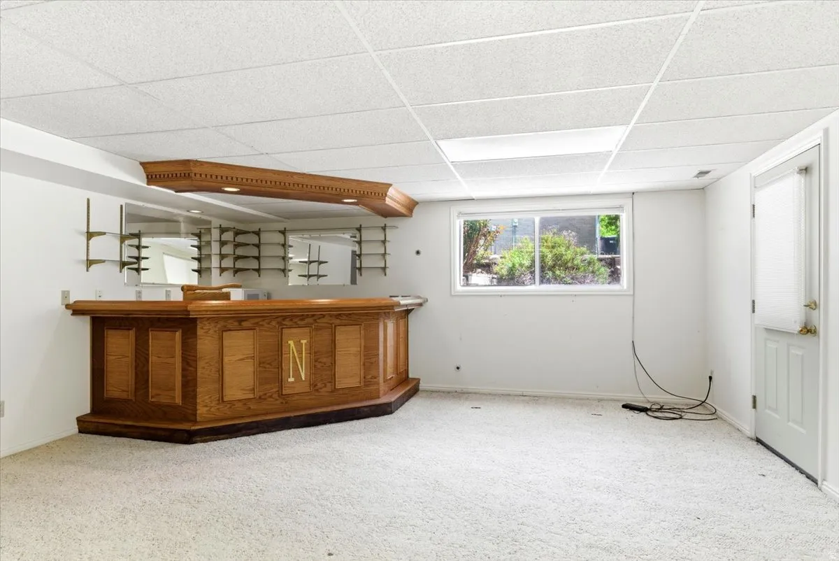 Built-in oak bar with mirrored backsplash and display shelving, recessed lighting, and a large window with exterior foliage