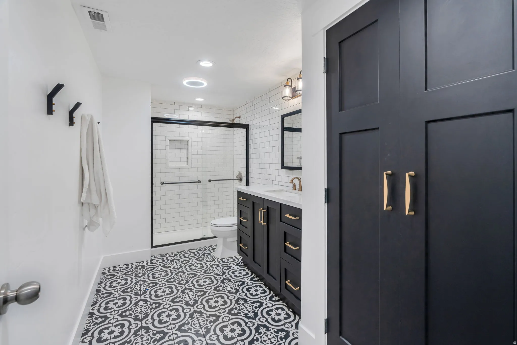 Bathroom featuring patterned floor tiles, a glass-enclosed shower with subway tile surround, a dark wood-finish vanity with a light-toned countertop, and gold-finish hardware