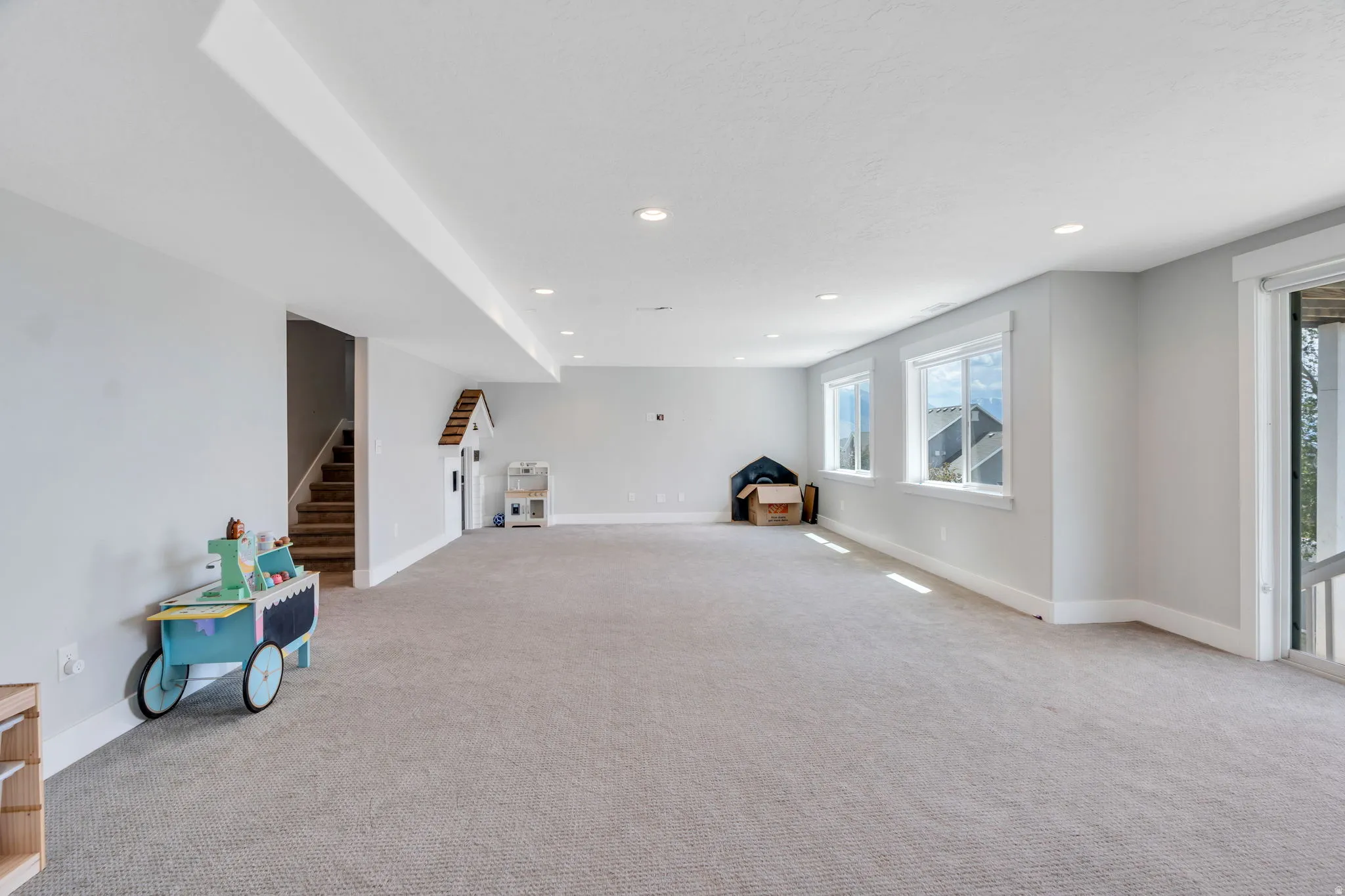 Expansive lower-level room featuring light gray carpeting, recessed lighting, multiple windows with white trim, a glass sliding door, and a permanent staircase with wood-finish treads