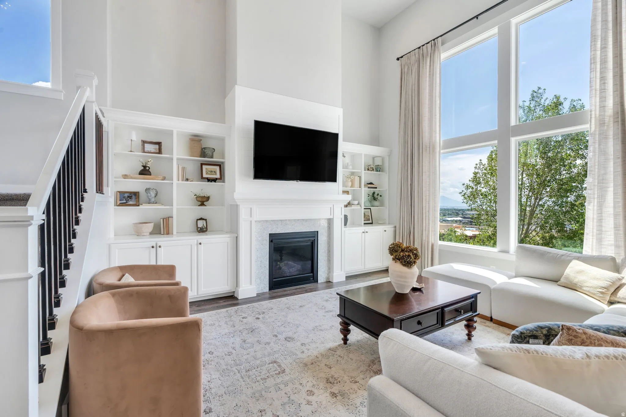 Two-story living area featuring a fireplace with a stone surround, built-in cabinetry, wood-finish flooring, and large windows with transom panels