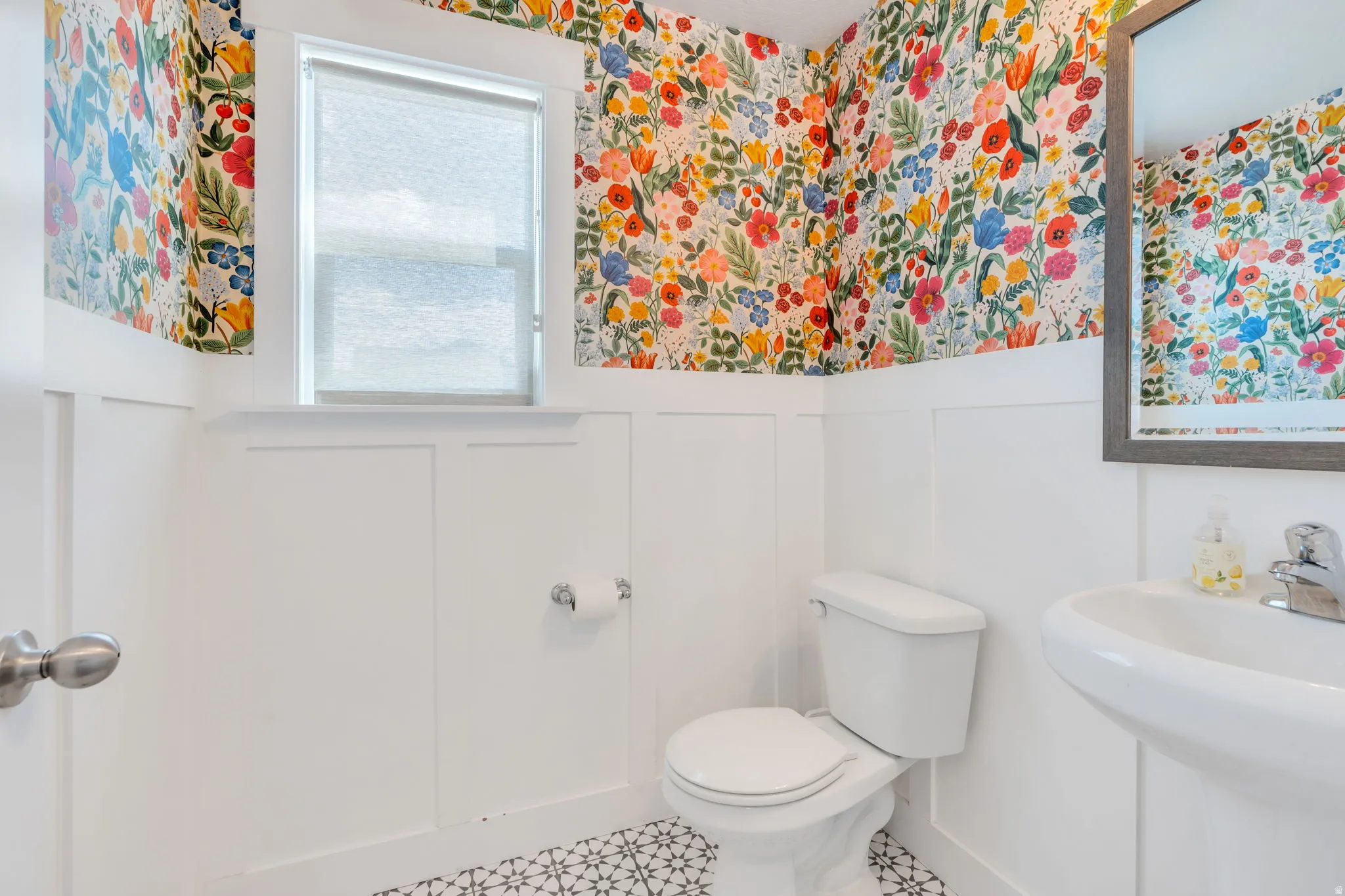 Bathroom featuring vibrant floral wallpaper, white wainscoting, a pedestal sink with a chrome faucet, and patterned floor tiles