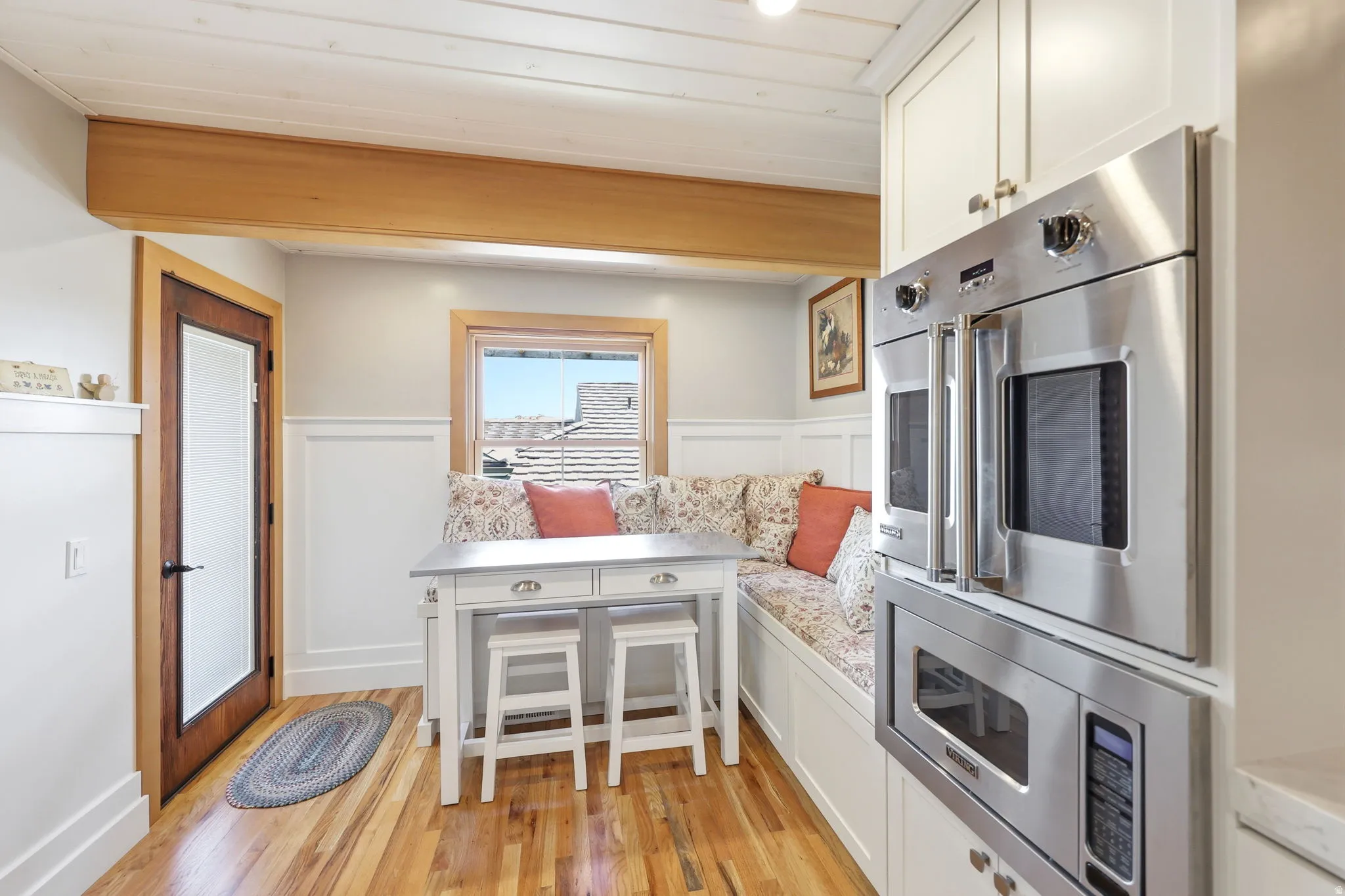 Built-in stainless steel double ovens and microwave, white shaker cabinetry, wood-finish flooring, exposed ceiling beam, and wainscoting
