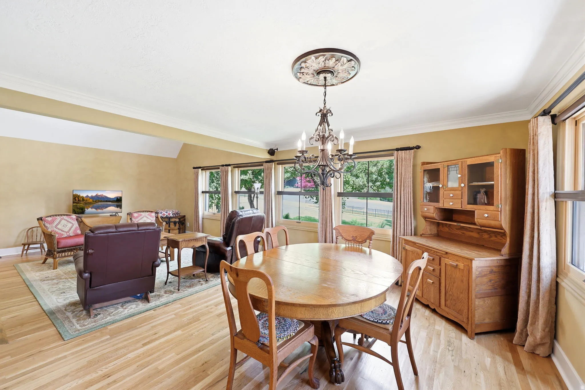 Open-concept living and dining area featuring wood-finish flooring, multiple windows, a decorative ceiling medallion with a chandelier, and crown molding