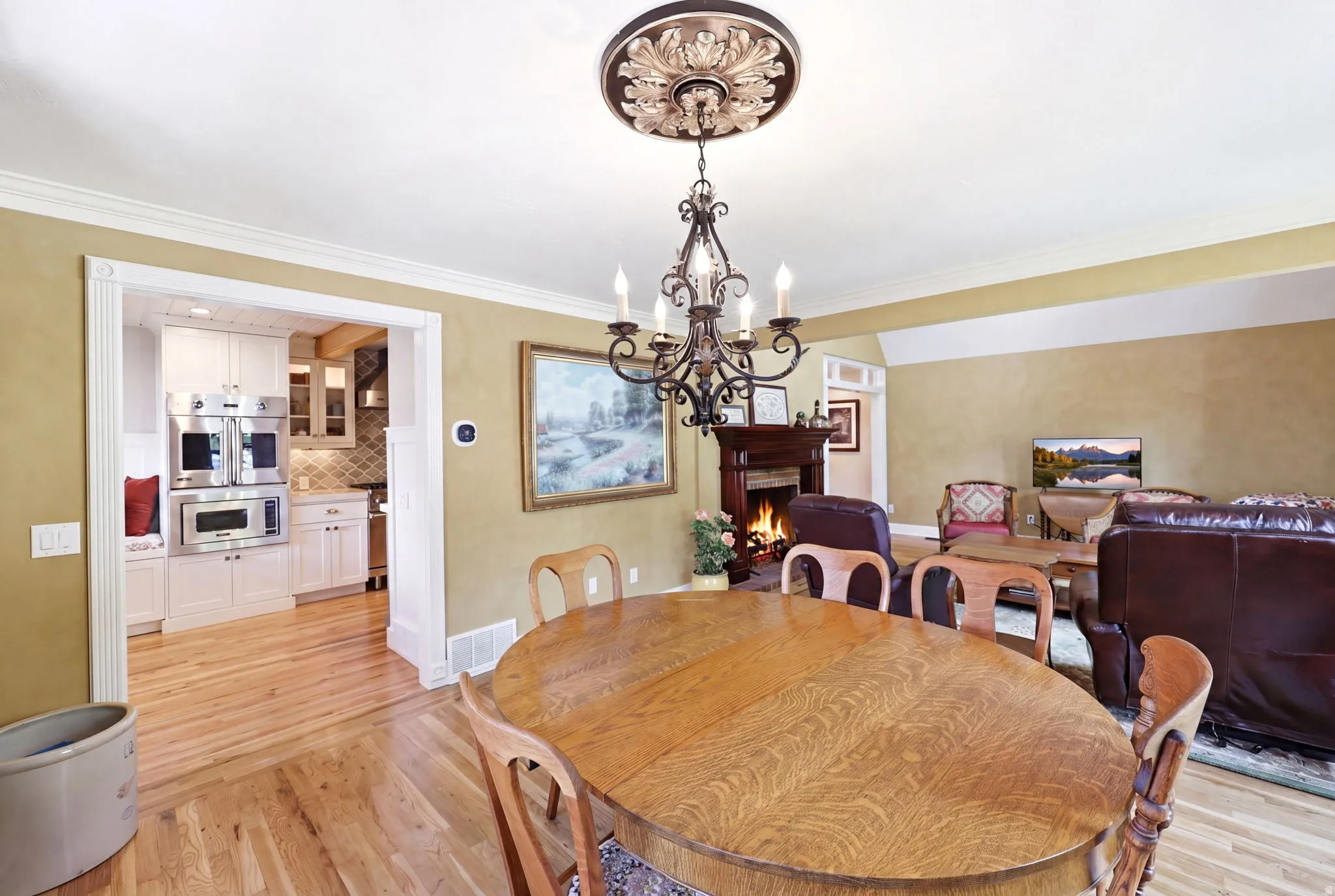Open-concept dining area featuring a decorative chandelier with an ornate ceiling medallion, wood-finish flooring, and a cased opening revealing a kitchen with stainless steel appliances