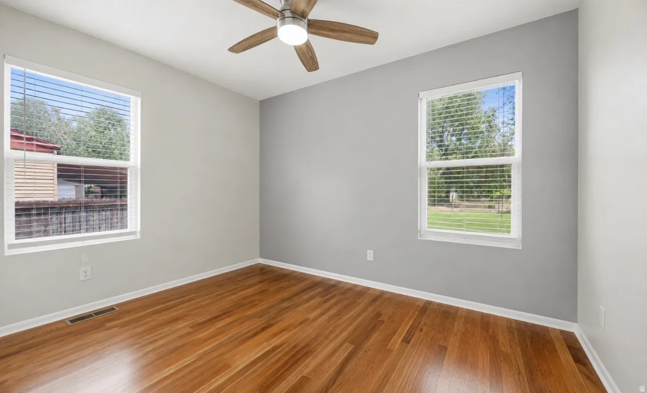 Room featuring wood-finish flooring, two windows with blinds, a ceiling fan with light fixture, and light gray painted walls