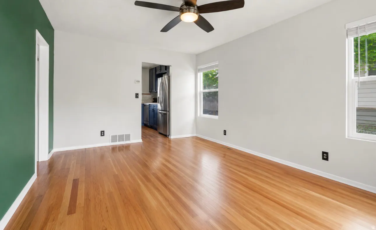 Spacious room featuring wood-finish flooring, light-toned walls, a dark-toned ceiling fan, and multiple windows
