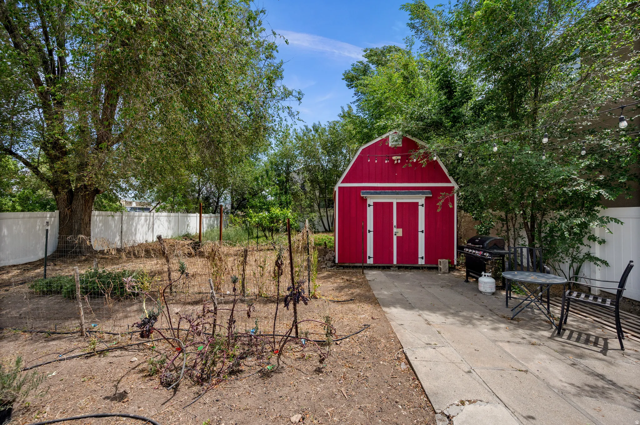Expansive backyard featuring a bright red storage shed with white trim, a concrete patio, mature trees, and a garden area