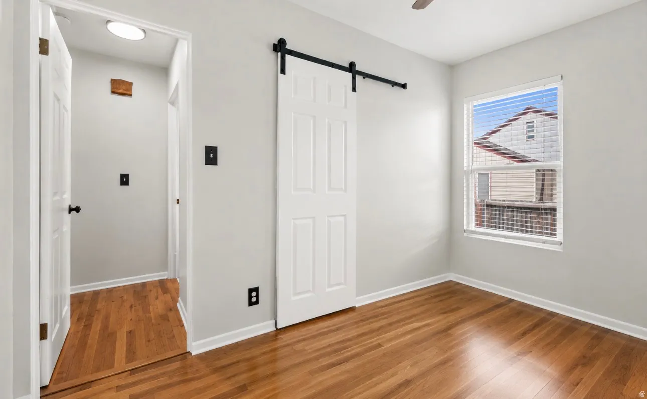 Interior room featuring wood-finish flooring, a white paneled barn door on a black track, a white window with horizontal blinds, and light gray wall paint