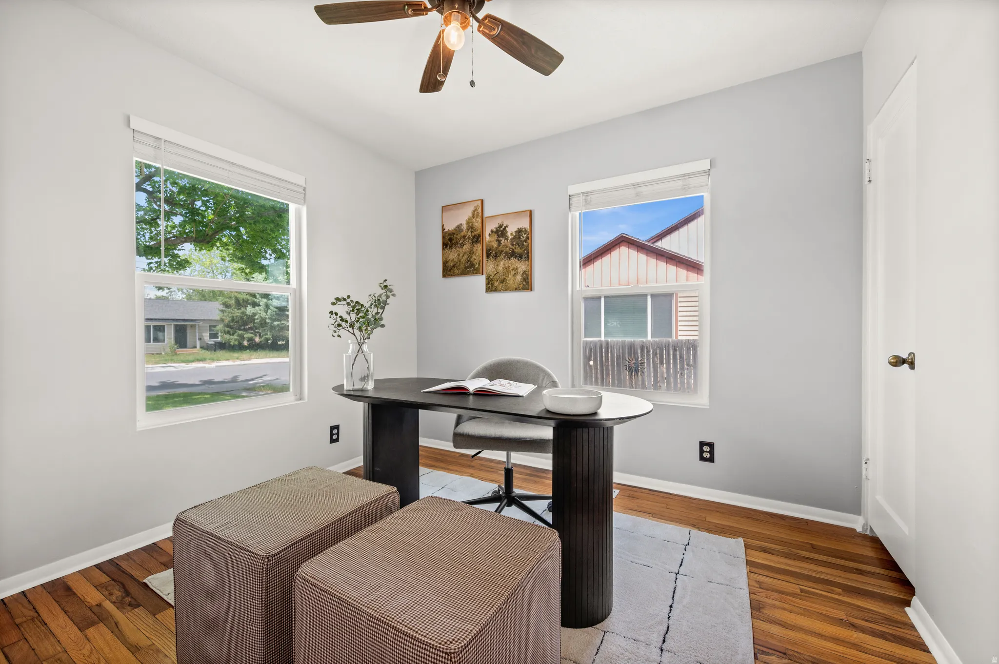 Bright room featuring two windows, wood-finish flooring, a ceiling fan with integrated lighting, and a white paneled door with a brass-toned knob