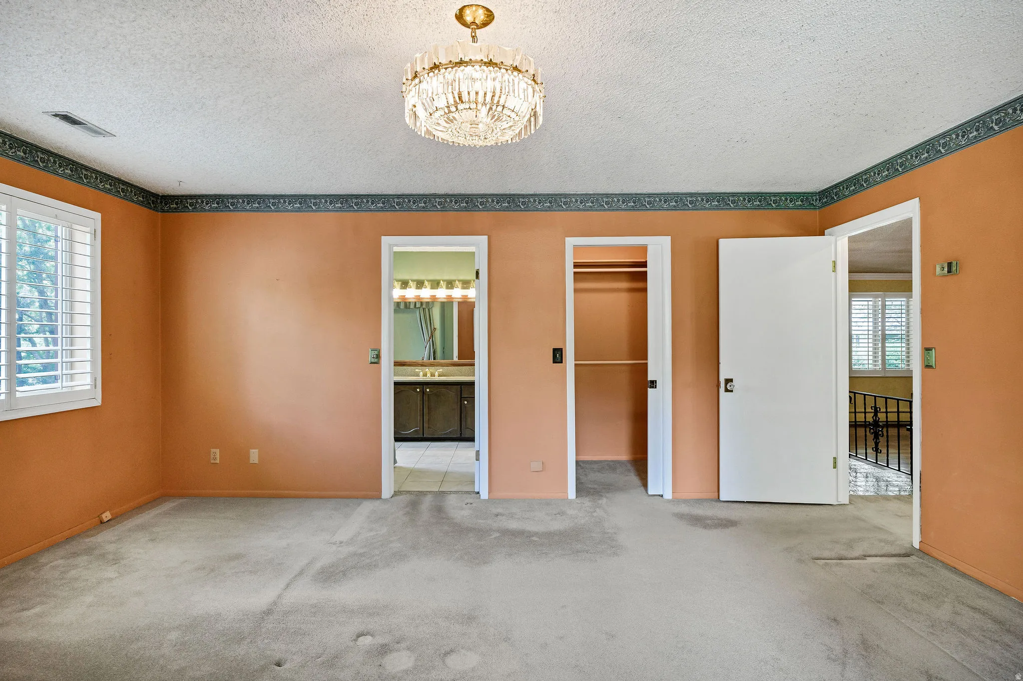 Carpeted interior room featuring a decorative chandelier, white plantation shutters, an open doorway revealing a bathroom vanity with multiple light fixtures, and a second open doorway showcasing a closet with shelving