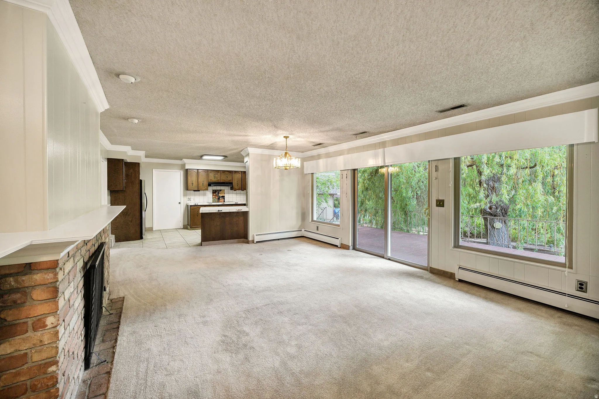 Open-concept living area featuring a brick fireplace with a white mantel, sliding glass doors, large windows, and an integrated kitchen with wood cabinetry