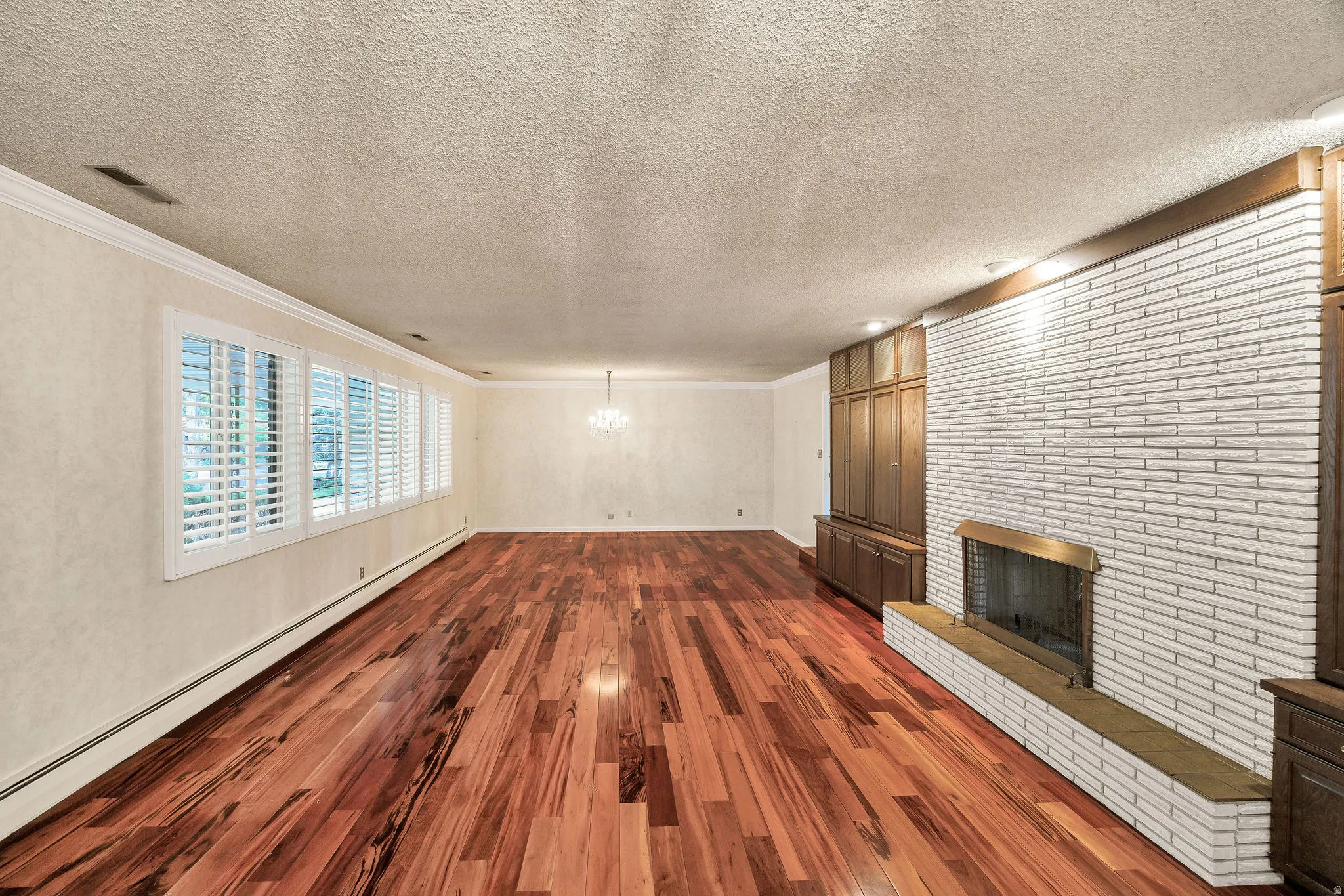 Spacious living area featuring polished wood-finish flooring, an extensive white brick fireplace with a brass-toned insert, built-in wood cabinetry, a row of windows with plantation shutters, and crown molding