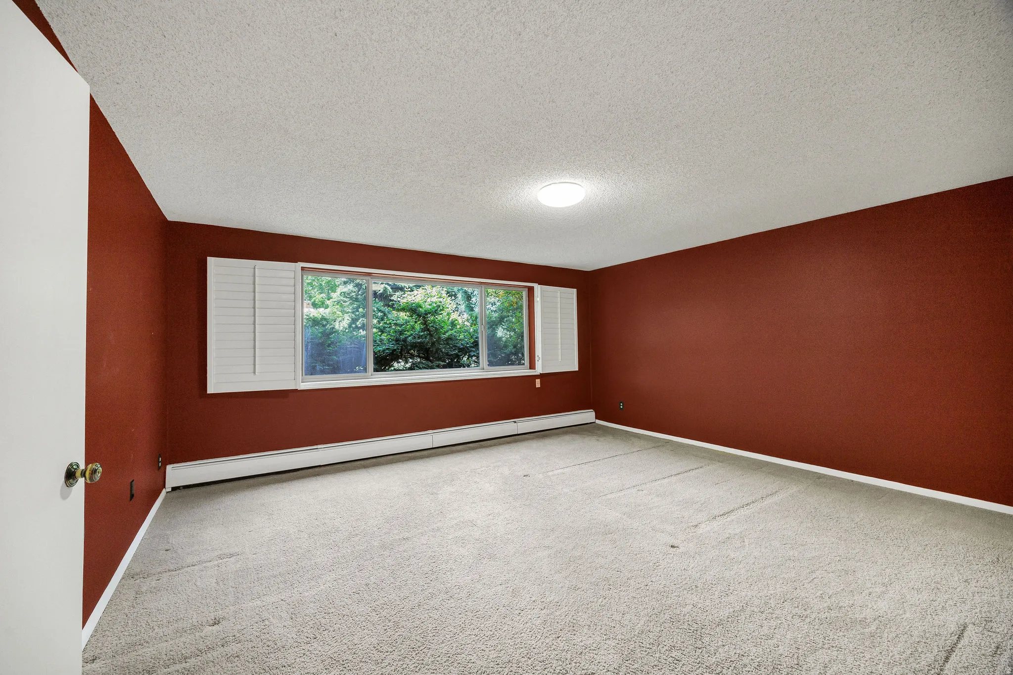 Spacious room featuring a wide window with white plantation shutters, light-toned carpeting, and baseboard heating