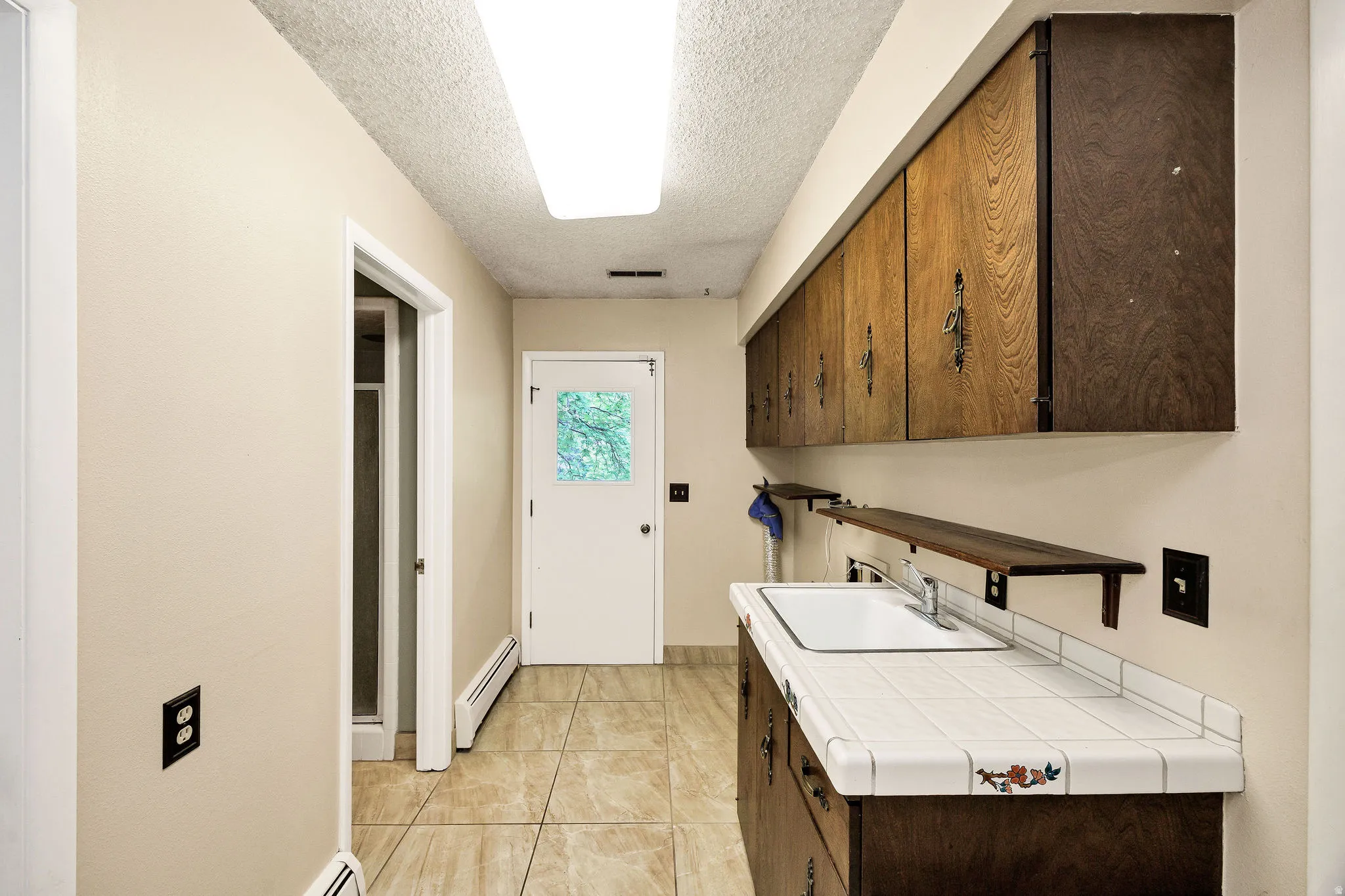Utility space featuring tile flooring, a white porcelain sink, tile countertops, wood-finish cabinetry, and a full-light exterior door