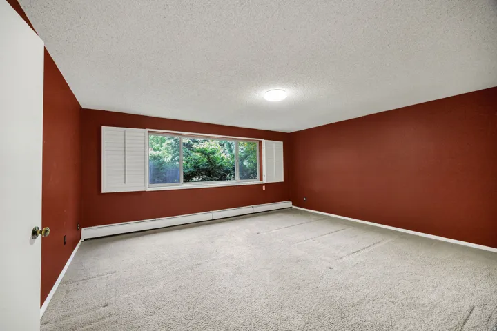 Spacious room featuring a wide window with white plantation shutters, light-toned carpeting, and baseboard heating