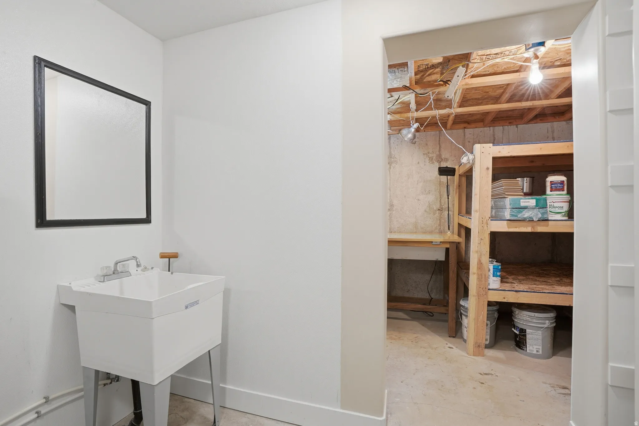 Storage and utility area featuring a white laundry sink with a chrome faucet, wall-mounted mirror, and exposed concrete flooring