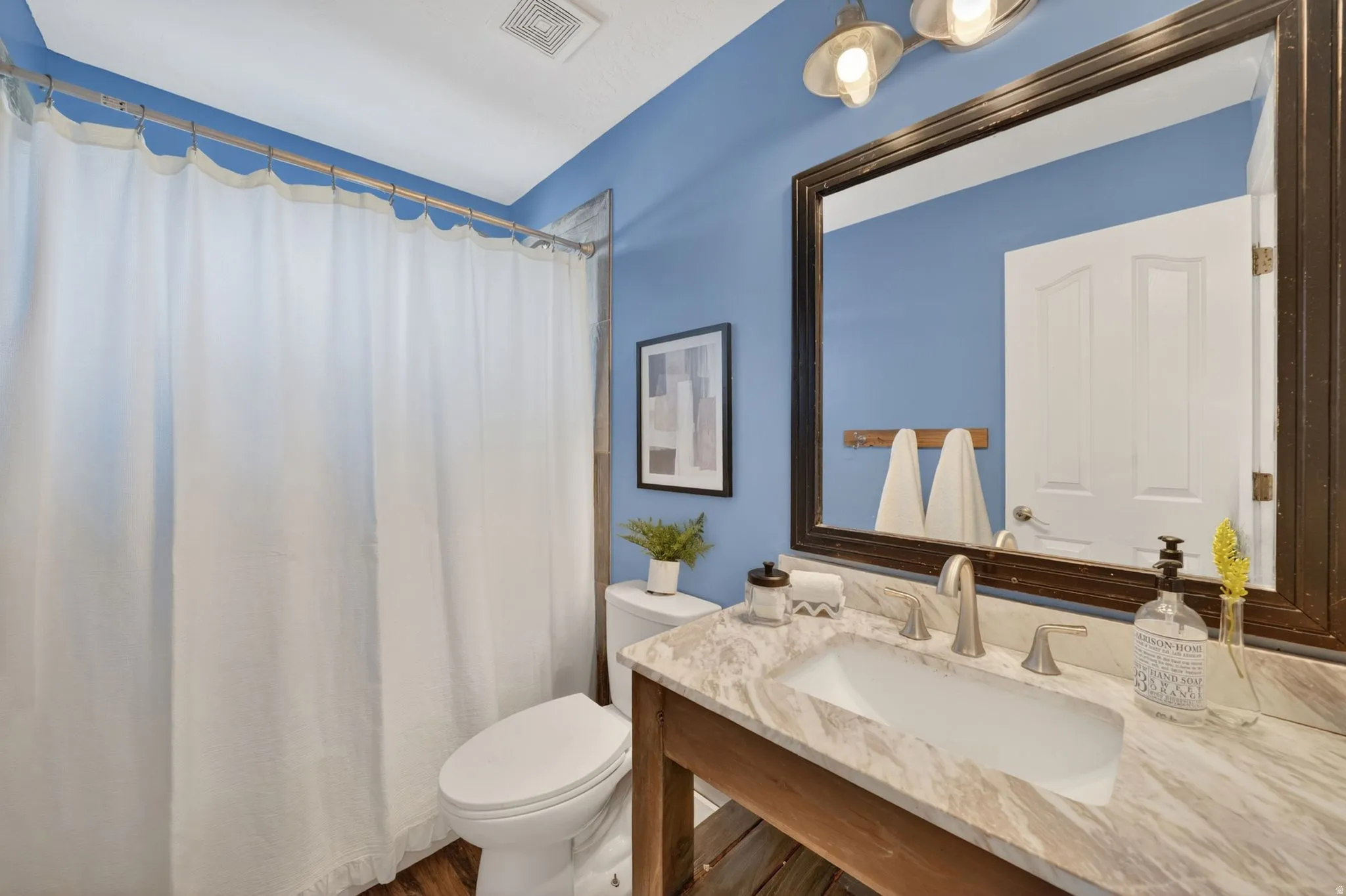 Bathroom featuring a rectangular undermount sink, a stone countertop, a dark wood-framed mirror, two-bulb vanity lighting, and wood-finish flooring
