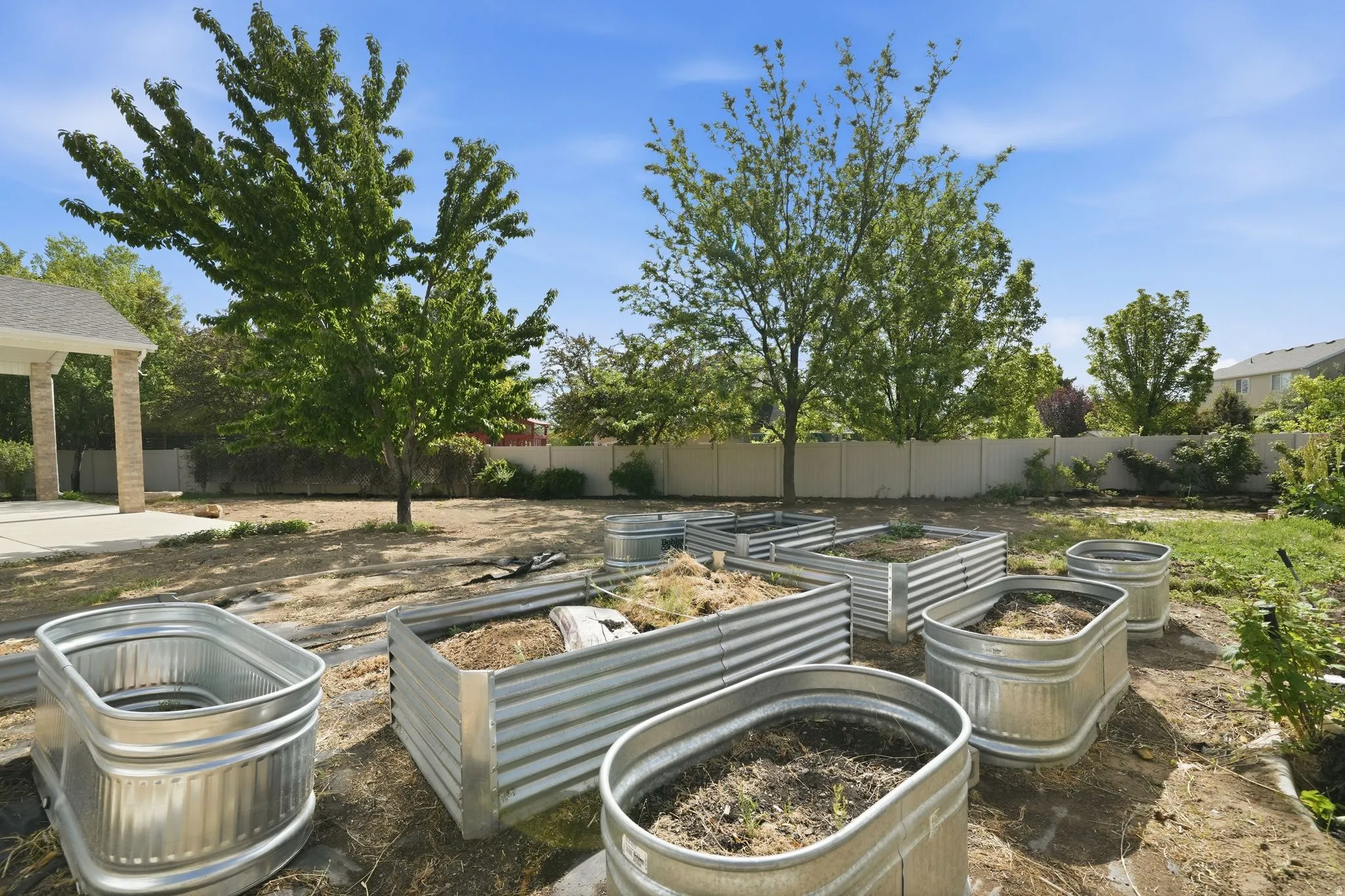 Expansive backyard featuring multiple corrugated metal raised garden beds, mature trees, and a concrete patio with a covered section