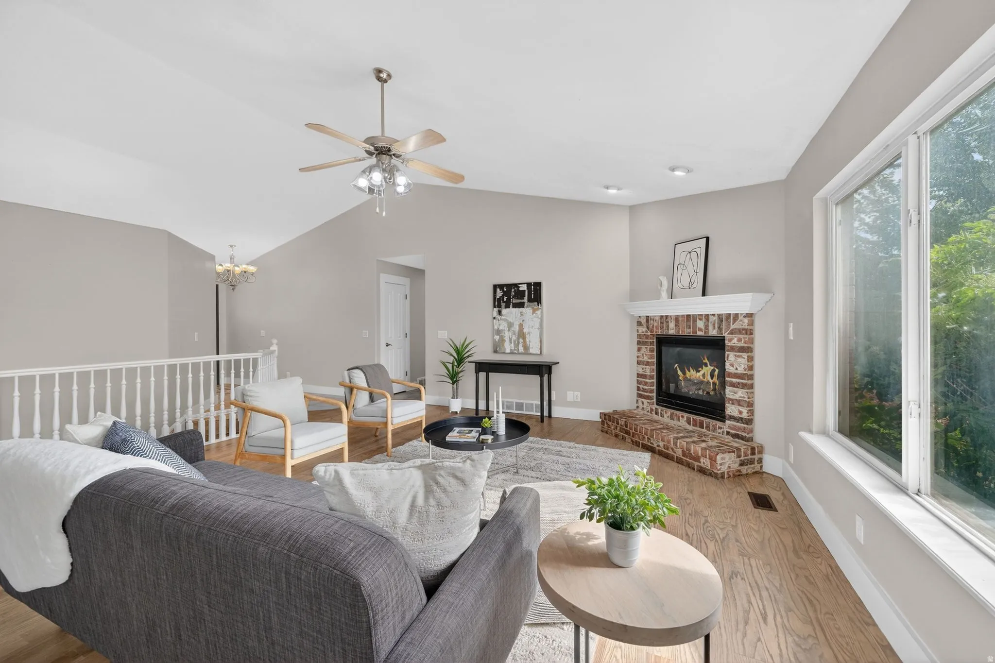 Living space featuring vaulted ceilings, wood-finish flooring, a brick fireplace with a white mantel, and large windows