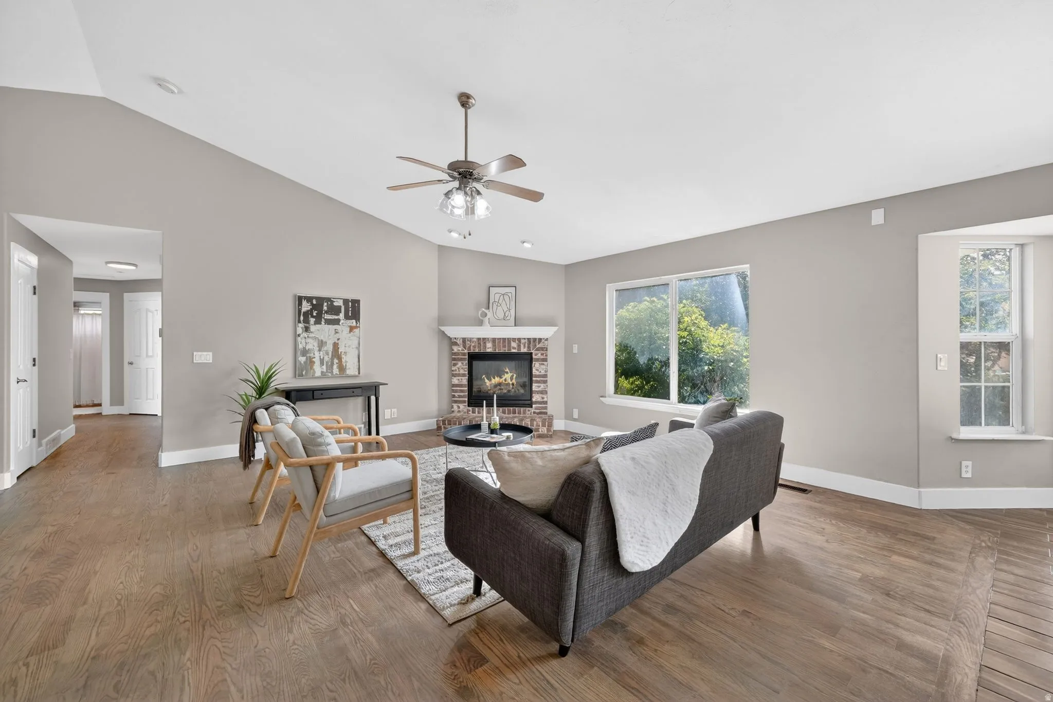 Spacious living area featuring vaulted ceilings, wood-finish flooring, a brick fireplace with a white mantel, and large windows