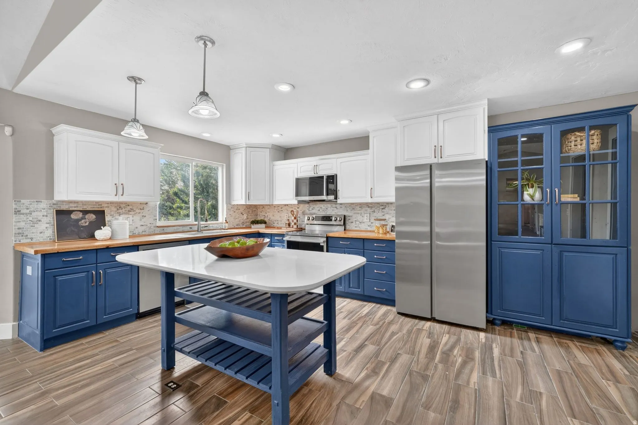 Kitchen featuring wood-finish flooring, two-tone cabinetry with white upper cabinets and blue lower cabinets, a blue kitchen island with a white countertop, wood-finish countertops, and a full-height pantry cabinet with glass doors