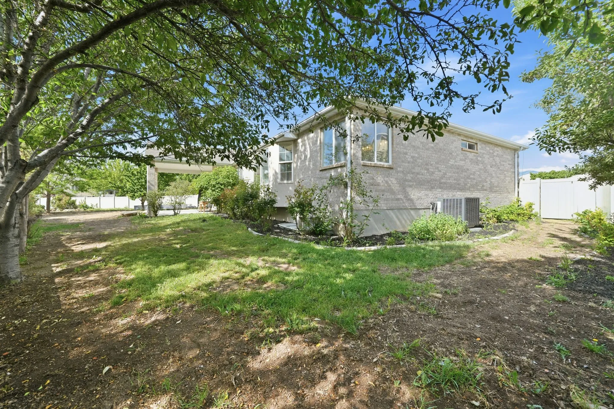 Expansive backyard featuring a partial grass area, mature trees, a white perimeter fence, and a paved side access