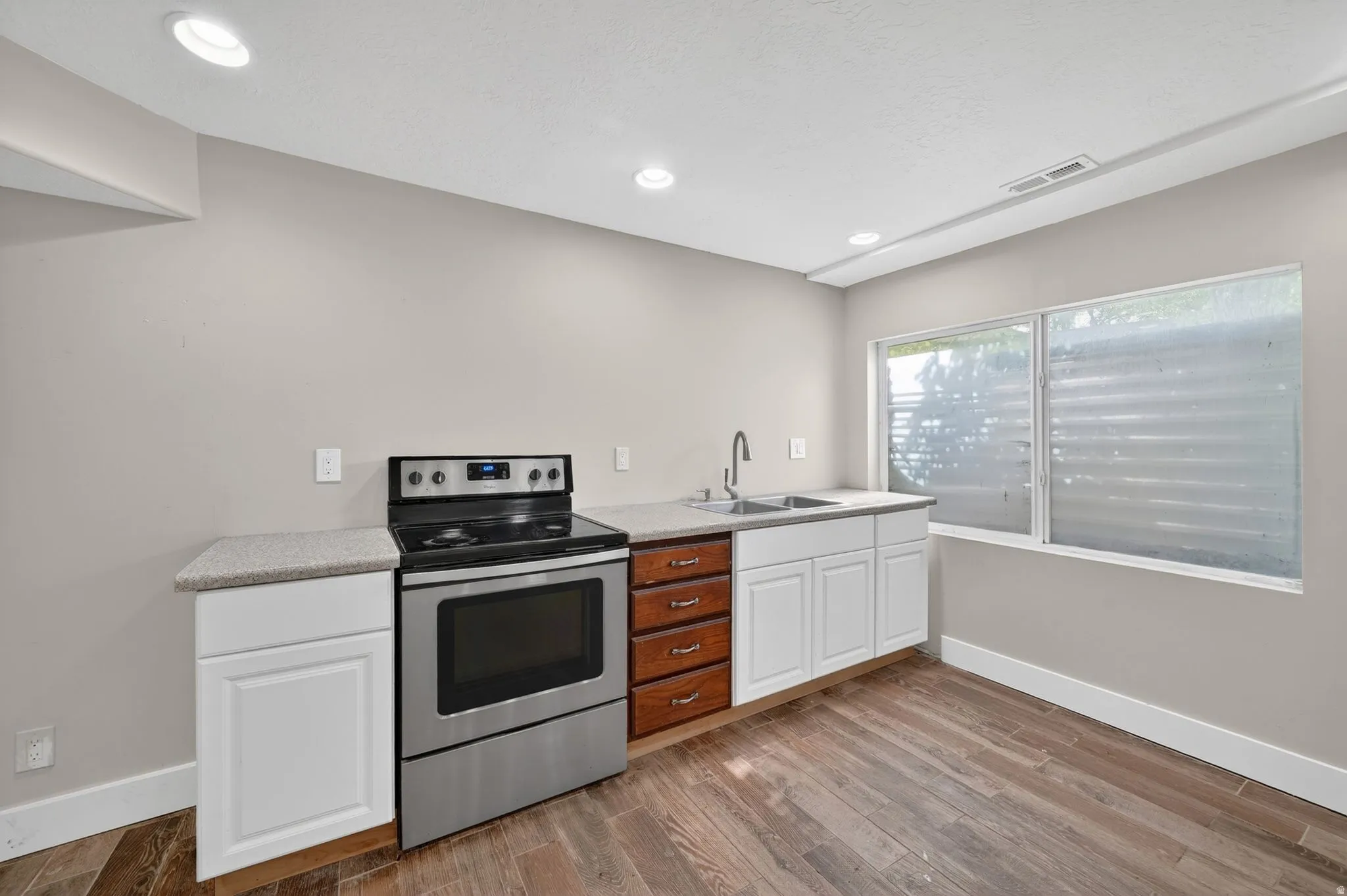 Compact kitchen featuring a stainless steel range, dual basin sink, light-toned countertops, mixed white and wood-finish cabinetry, and wood-finish flooring