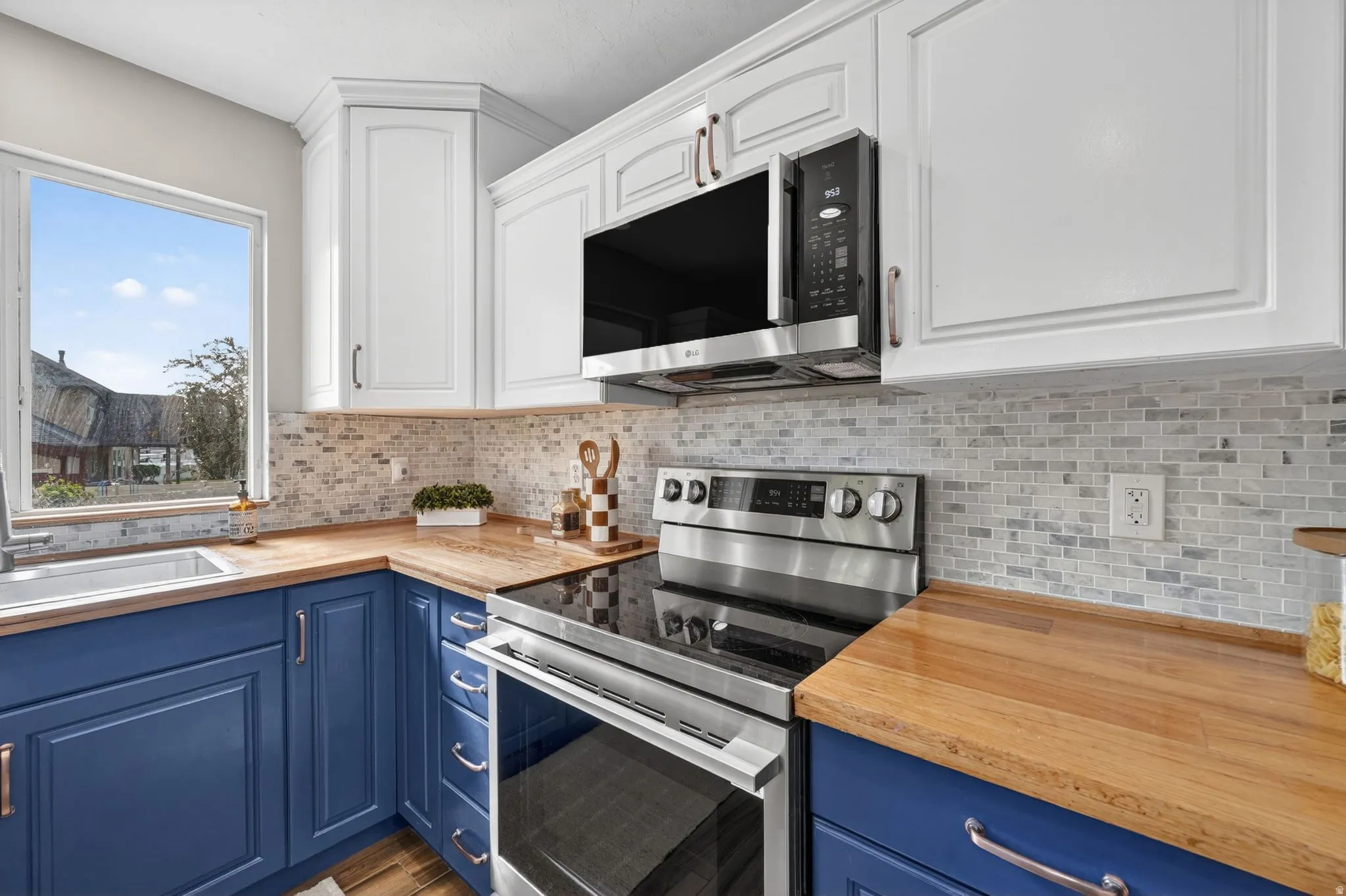 Kitchen featuring two-tone cabinetry with white upper and blue lower cabinets, a stainless steel electric range, an over-range microwave, wood-finish countertops, and a mosaic tile backsplash