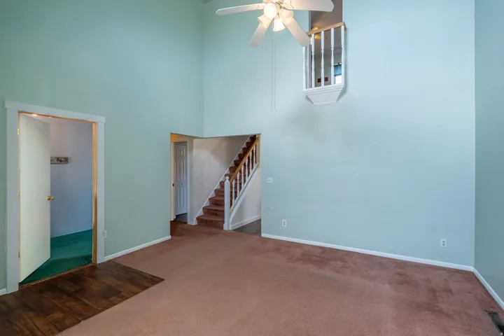 Spacious interior featuring a high vaulted ceiling with a ceiling fan, a prominent staircase with wood railing, an upper-level balcony, wood-finish flooring at the entrance, and light-colored carpeting throughout