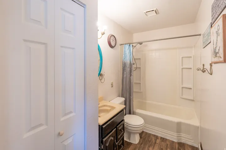 Full bathroom featuring a shower-tub combination with integrated shelving, dark wood vanity with a light-toned countertop, white toilet, and wood-finish flooring