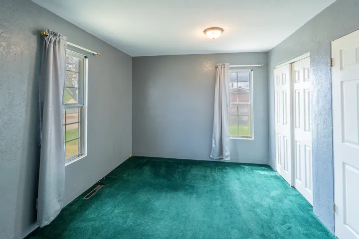 Room featuring plush green carpeting, two windows with white frames, a ceiling-mounted light fixture, and white bi-fold closet doors