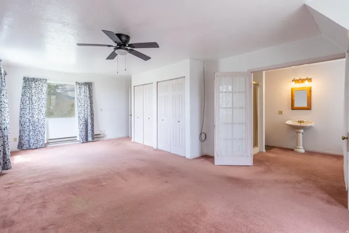 Spacious room featuring ceiling fan, three bi-fold closet doors, window with baseboard heating, and an adjacent bathroom with pedestal sink, mirror, and wall-mounted lighting