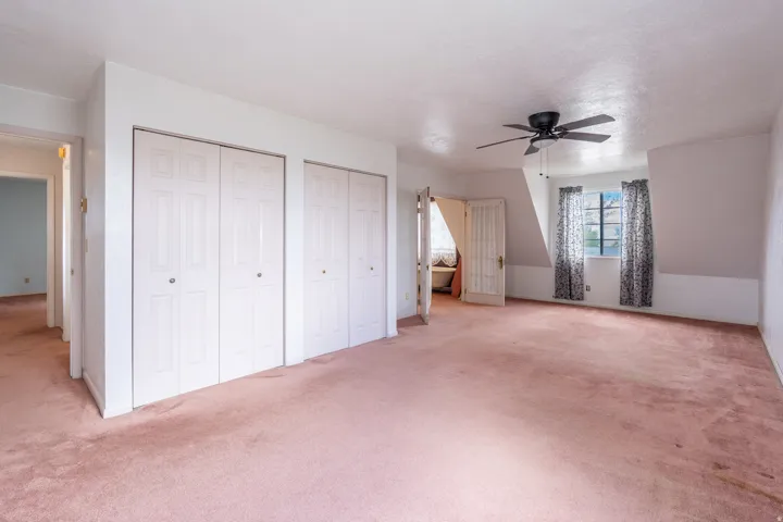 Spacious room featuring neutral carpeting, multiple bi-fold closet doors, a ceiling fan, and window with natural light