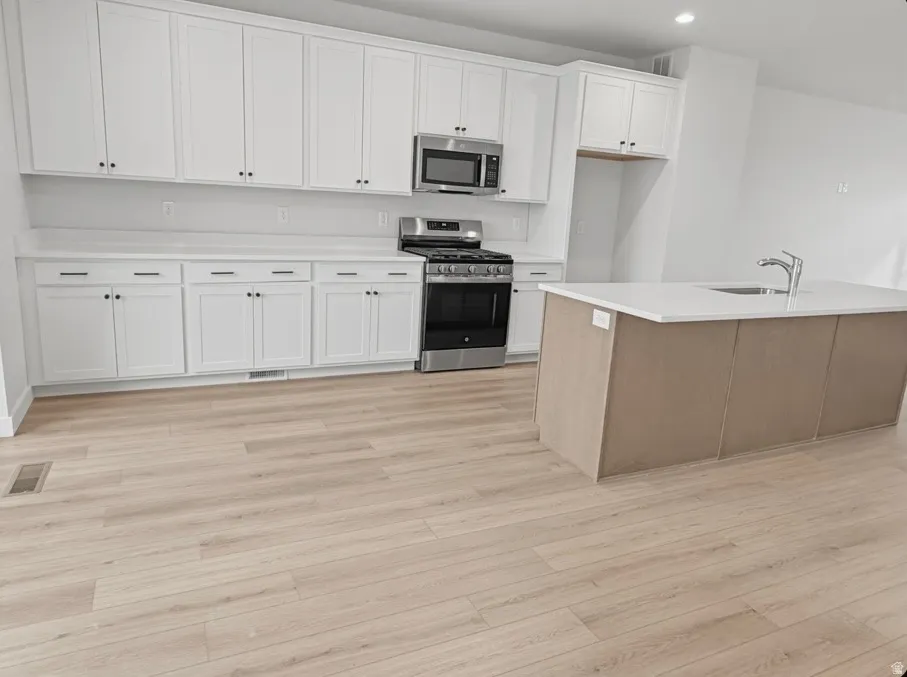 Modern kitchen featuring white shaker cabinetry, stainless steel appliances, light wood-finish flooring, a contemporary island with a built-in sink, and light-toned countertops