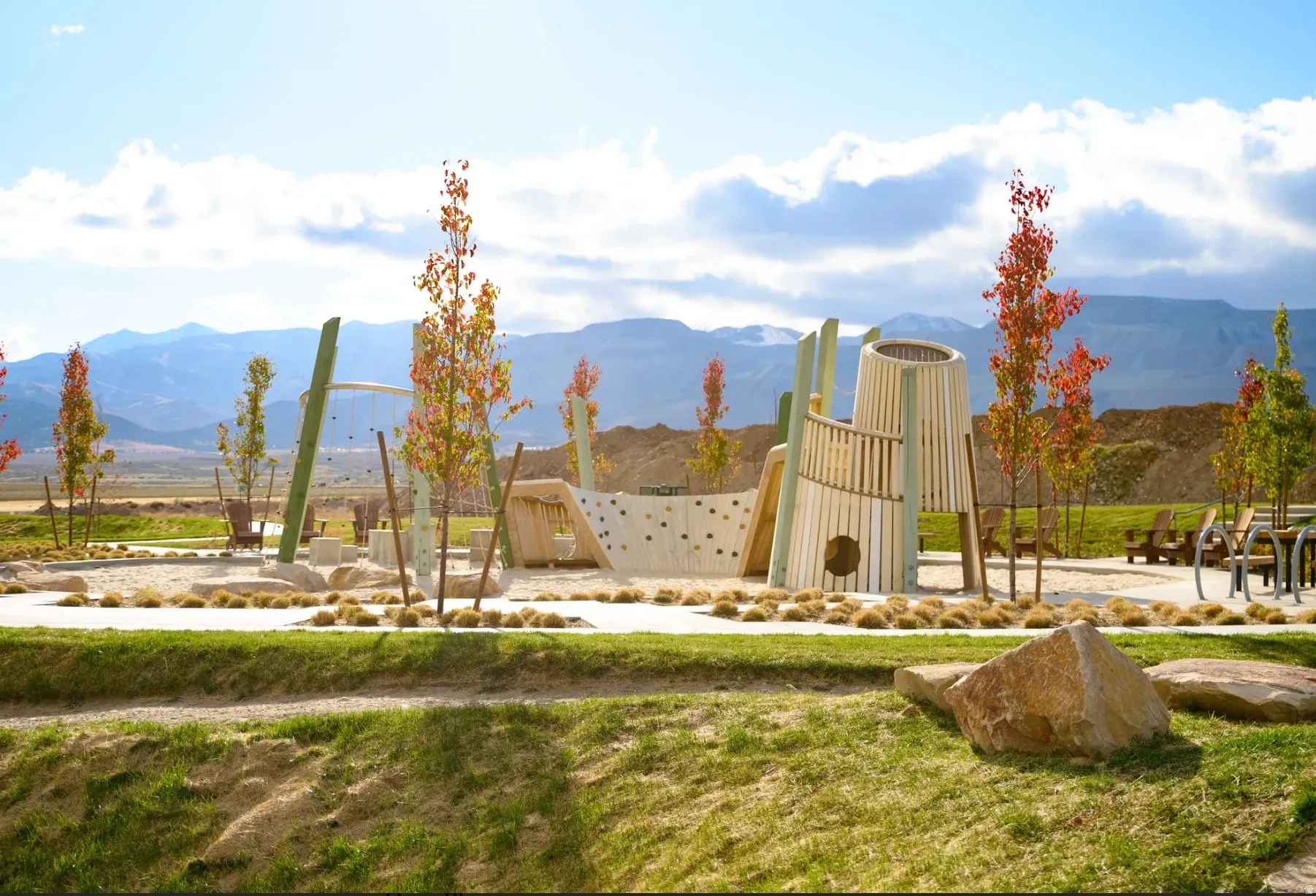 Community playground featuring modern wood and rock climbing structures, a sandy play area, integrated seating, and mature landscaping