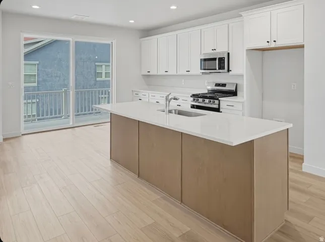 Modern kitchen featuring a large central island with an undermount sink, light wood-finish flooring, white shaker cabinetry, stainless steel appliances, and a sliding glass door opening to a private balcony