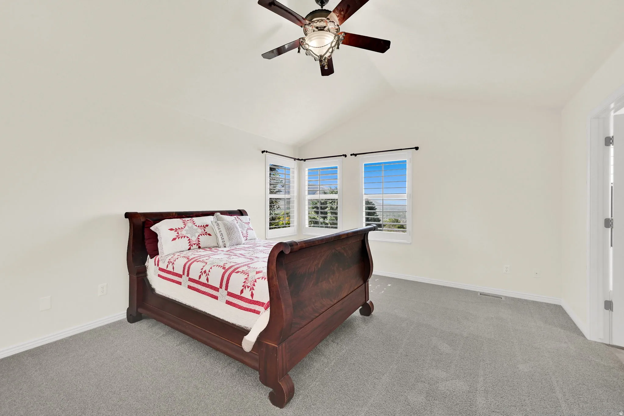 Vaulted ceiling with a ceiling fan, three windows with plantation shutters, light-colored walls, and gray carpet flooring