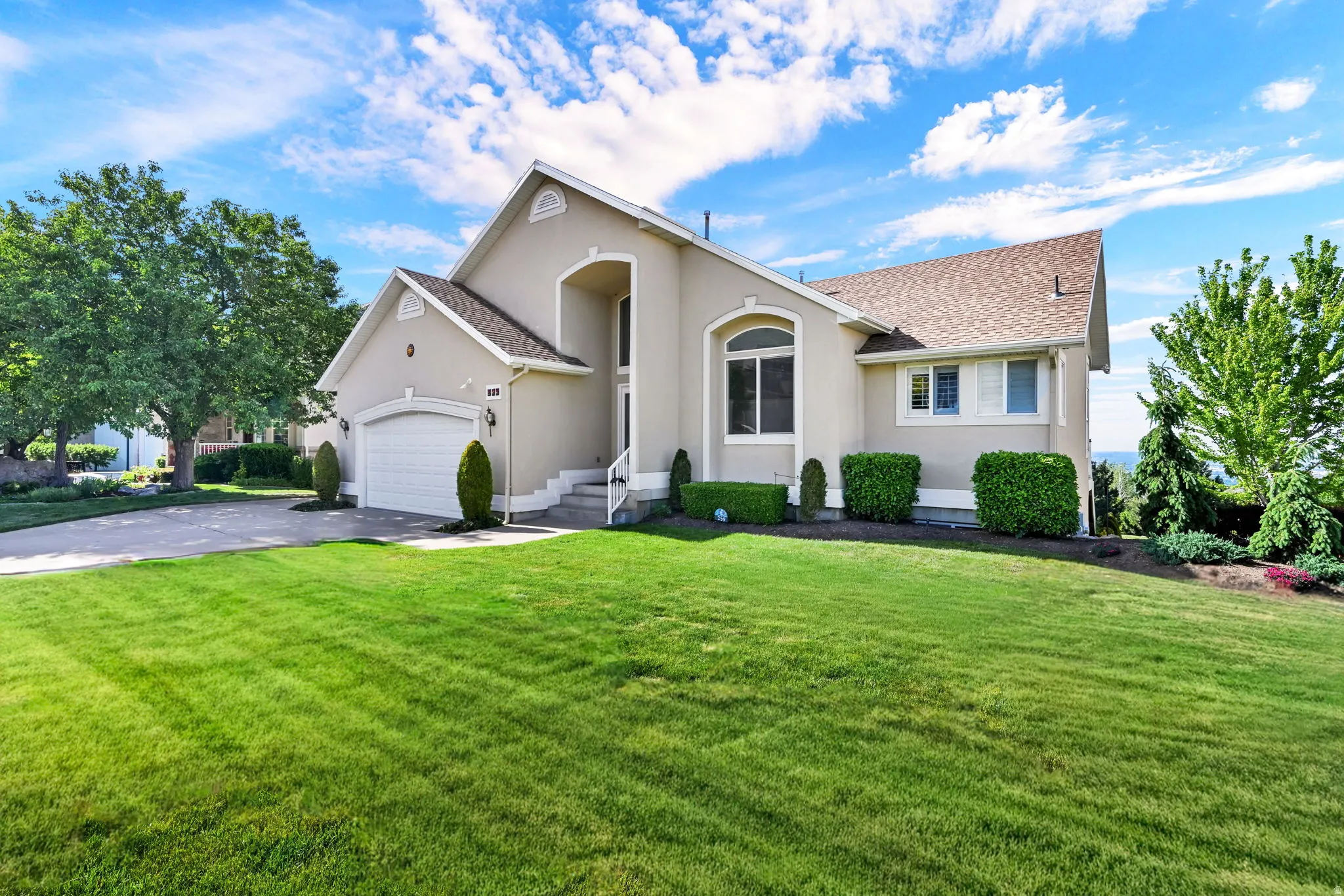 Stucco exterior residence featuring a two-car garage, pitched shingle roof, arched entryway with steps, and a landscaped front yard with a paved driveway
