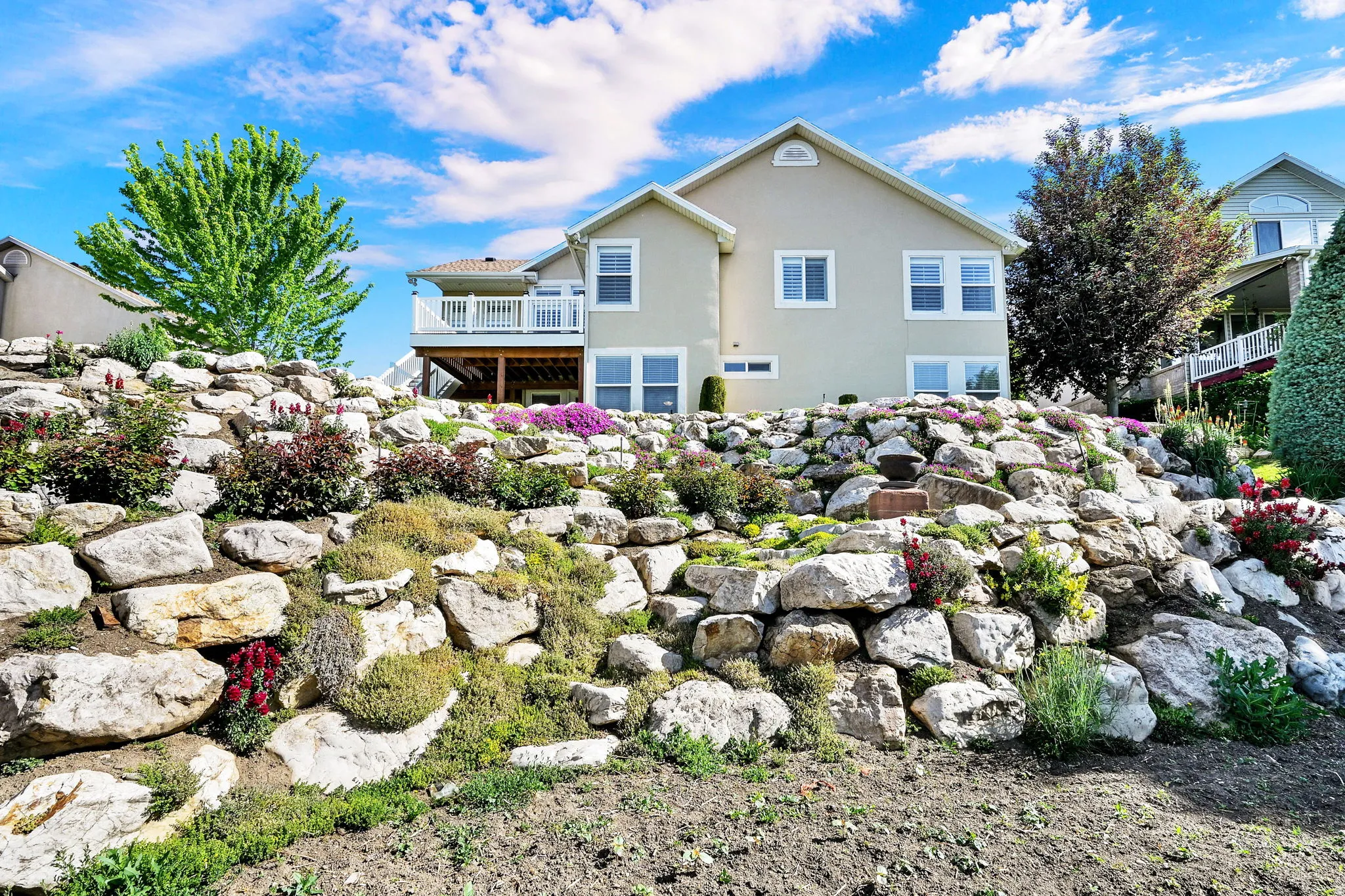 Rear exterior featuring a multi-level rock retaining wall with diverse plantings, a second-story balcony with white railing, and a stucco finish
