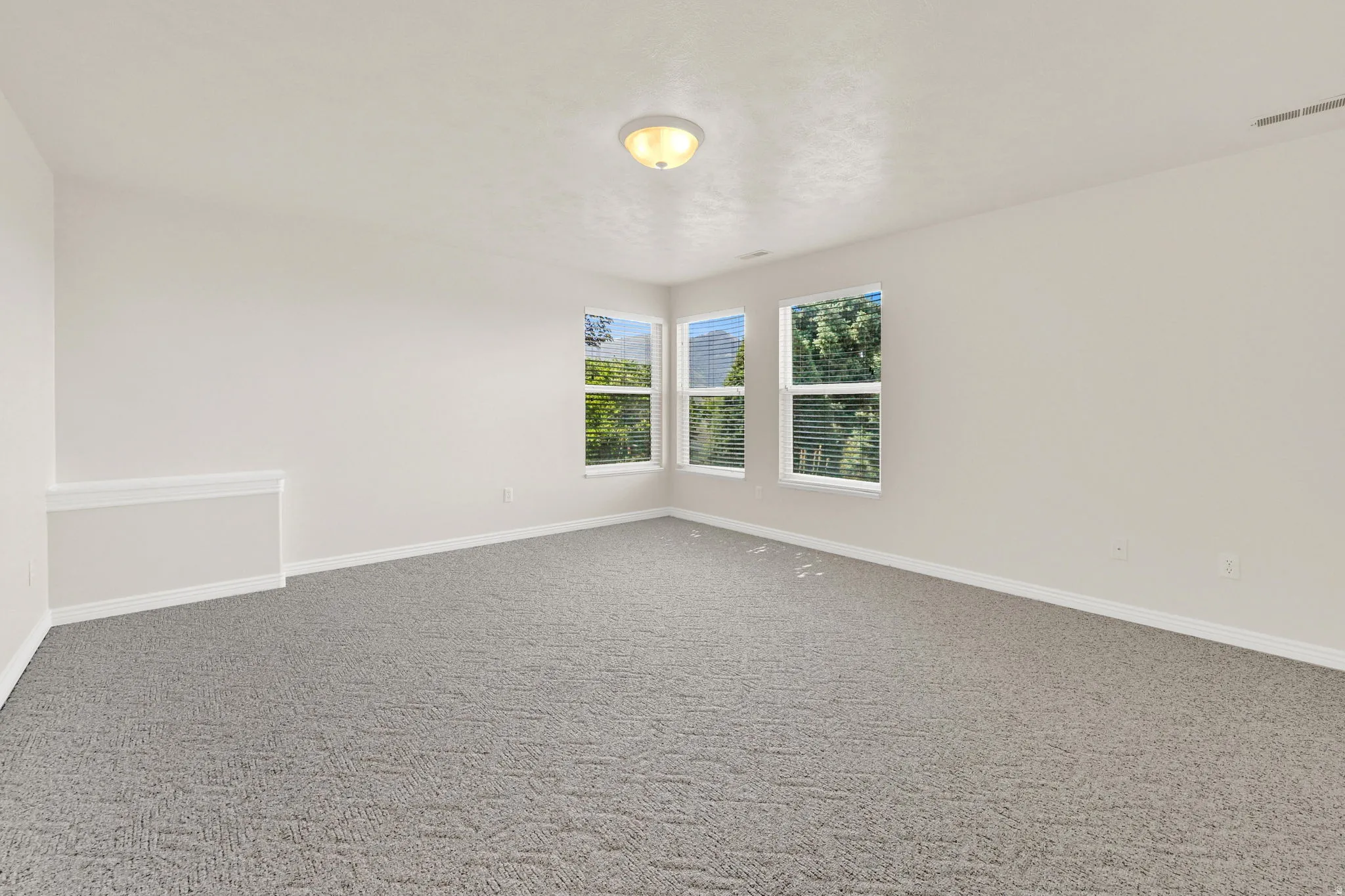 Spacious interior featuring three windows, neutral wall tones, and gray patterned carpeting