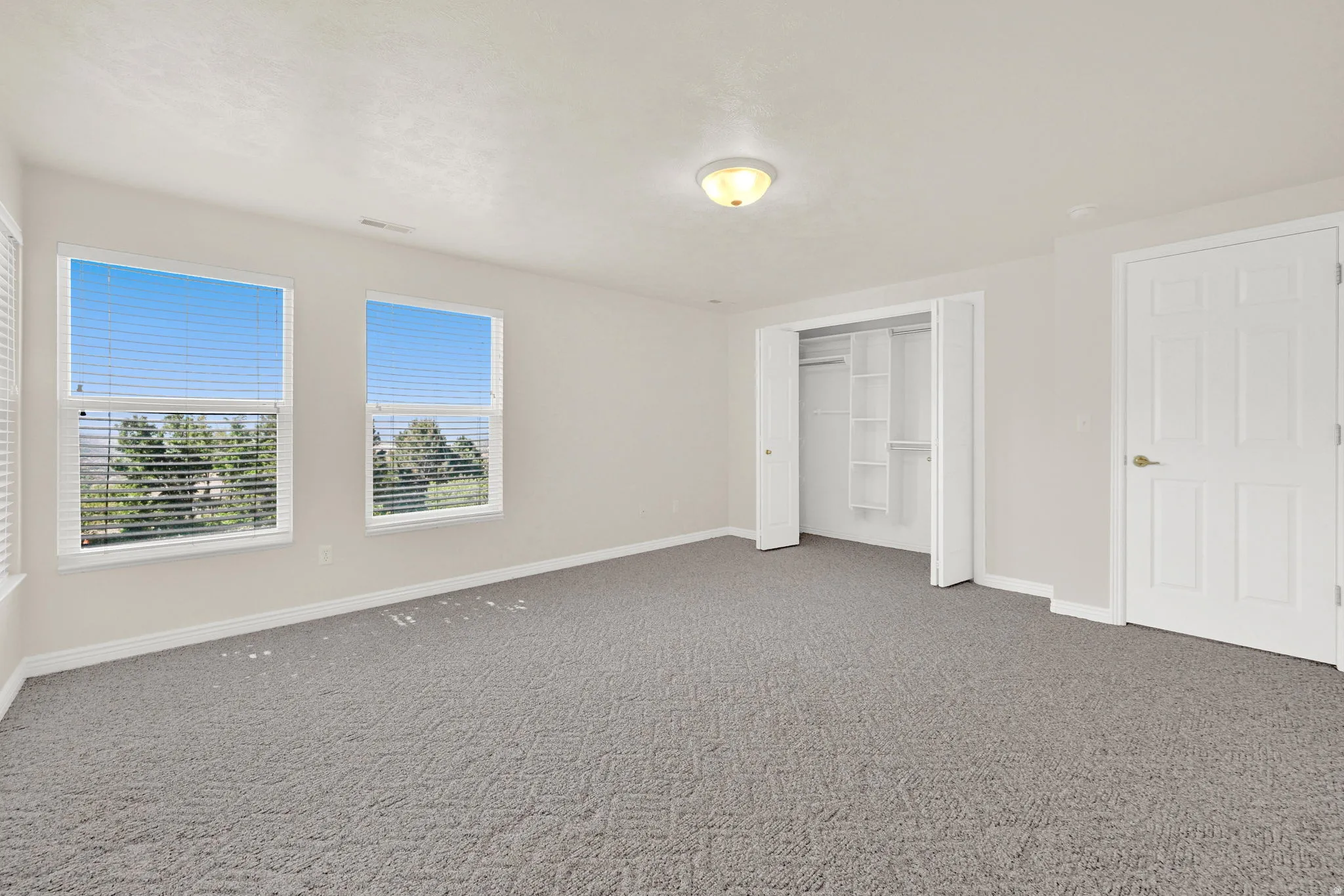 Spacious room with two large windows featuring horizontal blinds, neutral wall paint, gray patterned carpet, a built-in closet with shelving, and a six-panel door